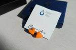Orange heart-shaped earrings on a card with 'Atelier Meer' branding.